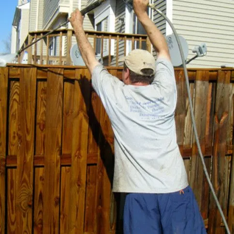 Crew member soft-washing a wood fence — back of head, branded grey shirt, pressure wand reaching up